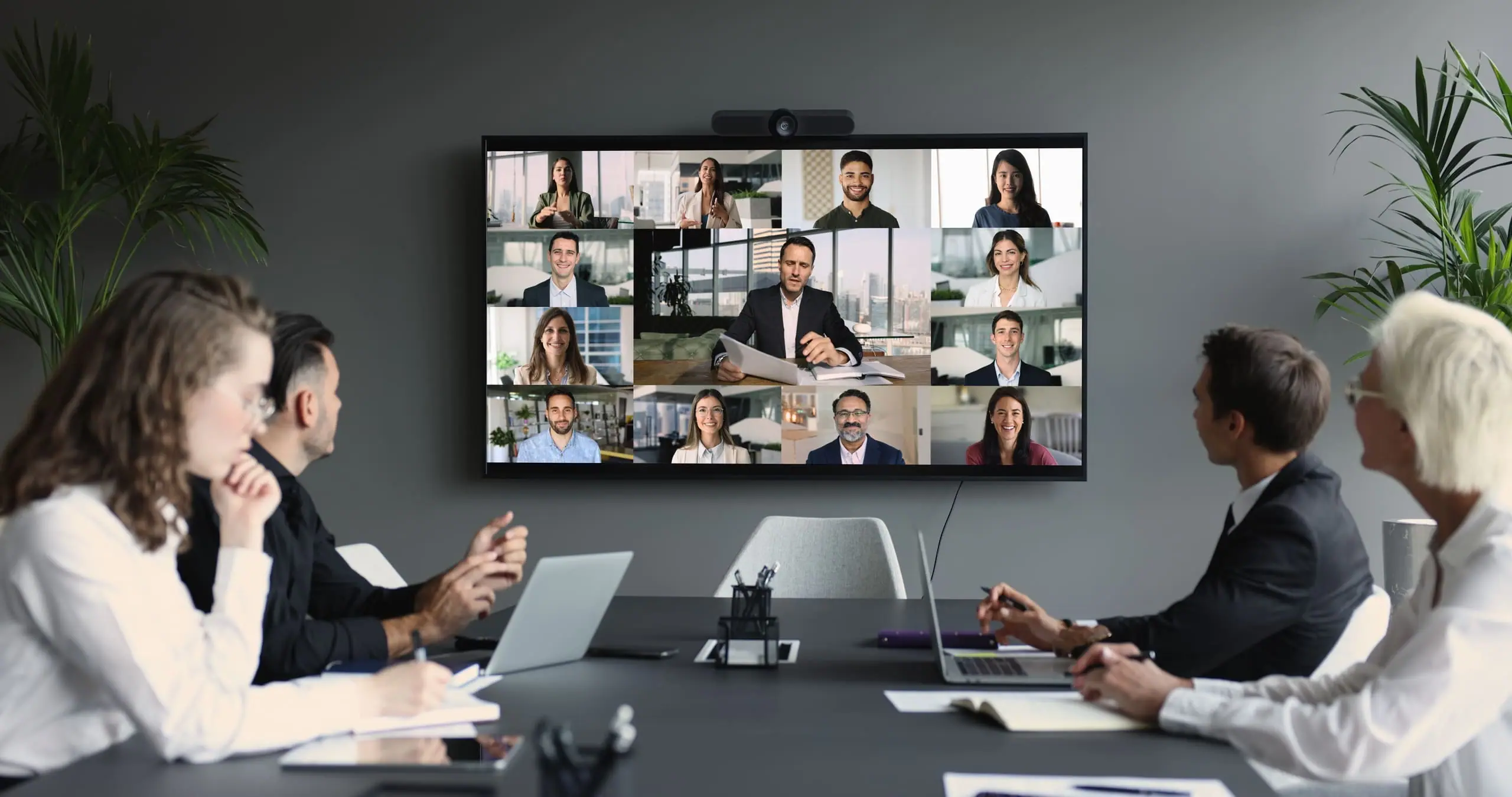 Colleagues around a boardroom table on a video call, with remote participants shown on a large wall screen.